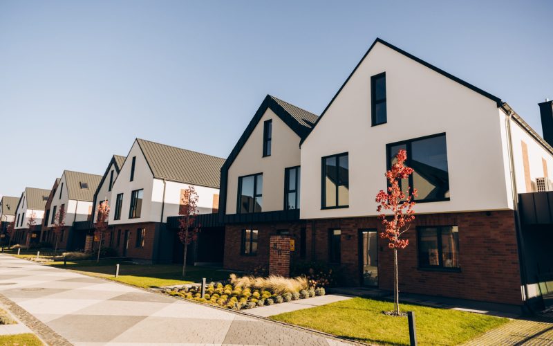 Row of contemporary white and brick townhouses in a newly developed residential neighborhood, featuring clean architectural lines, green lawns, and small autumn trees under a clear blue sky